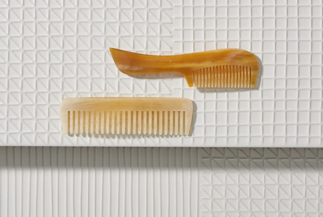 Two horn combs in brown and cream colors displayed on a textured white tile background.