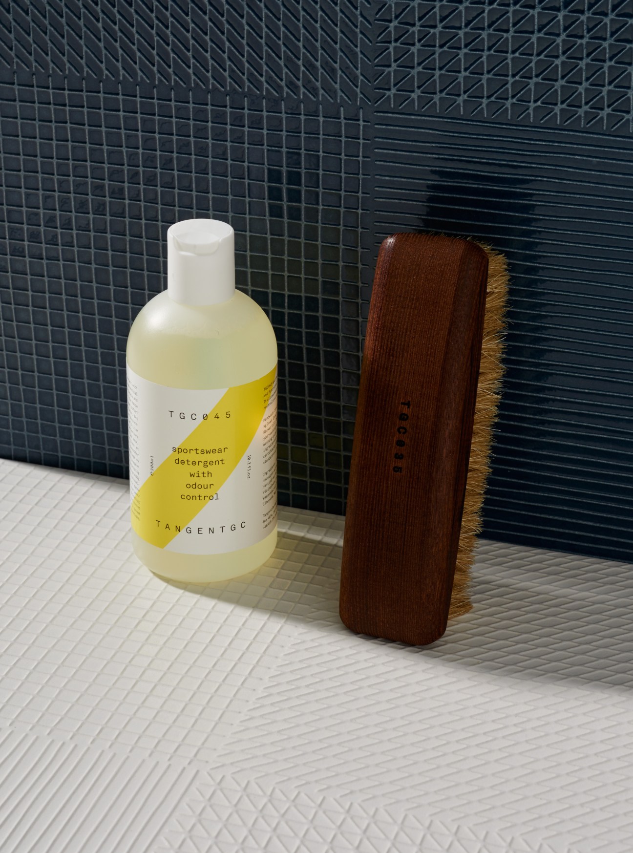 Tangent Garment Care sportswear detergent bottle and wooden shoe brush on tiled bathroom surface.
