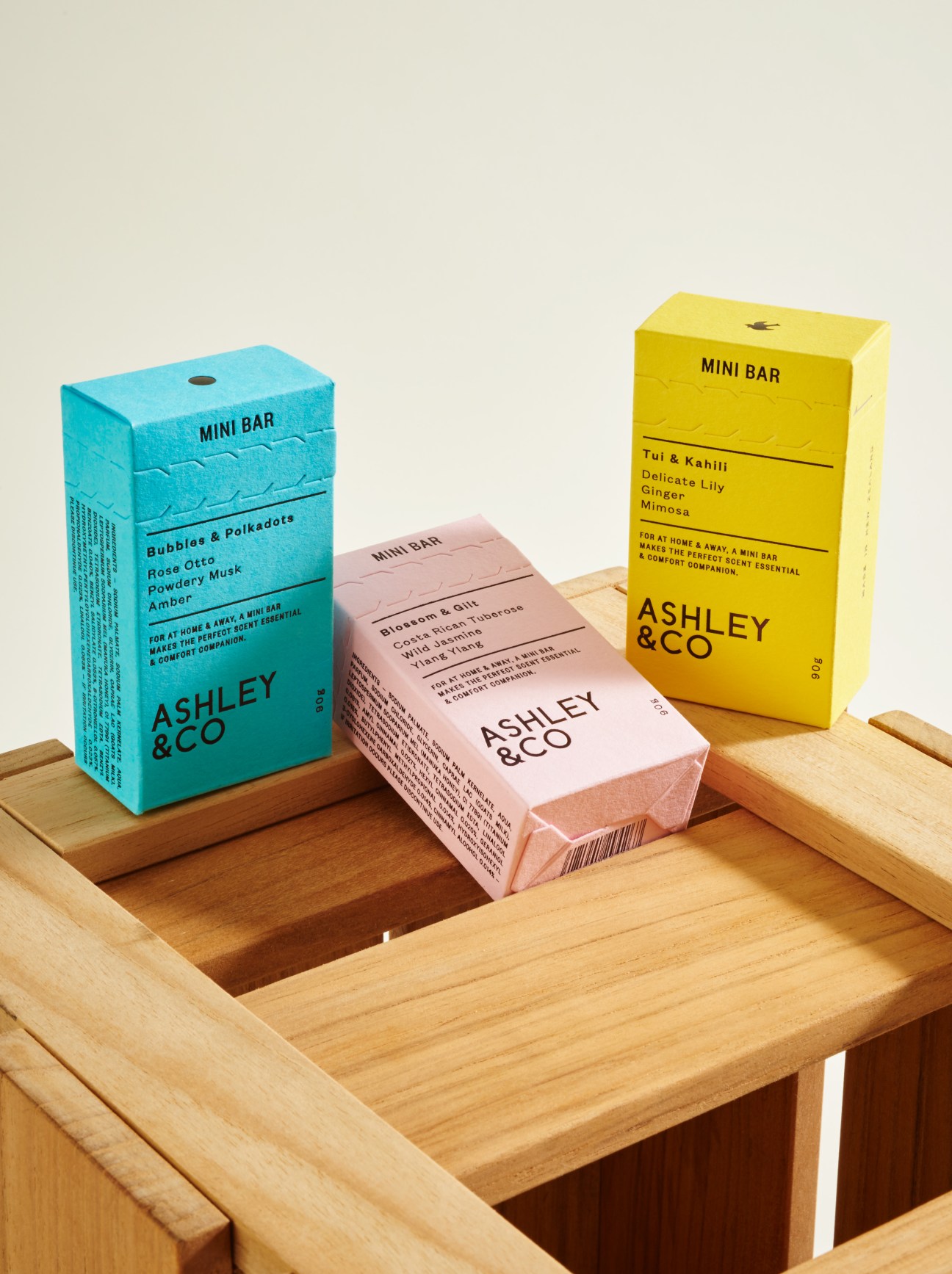 Three colorful Mini Bar soap boxes by Ashley & Co displayed in a wooden tray.