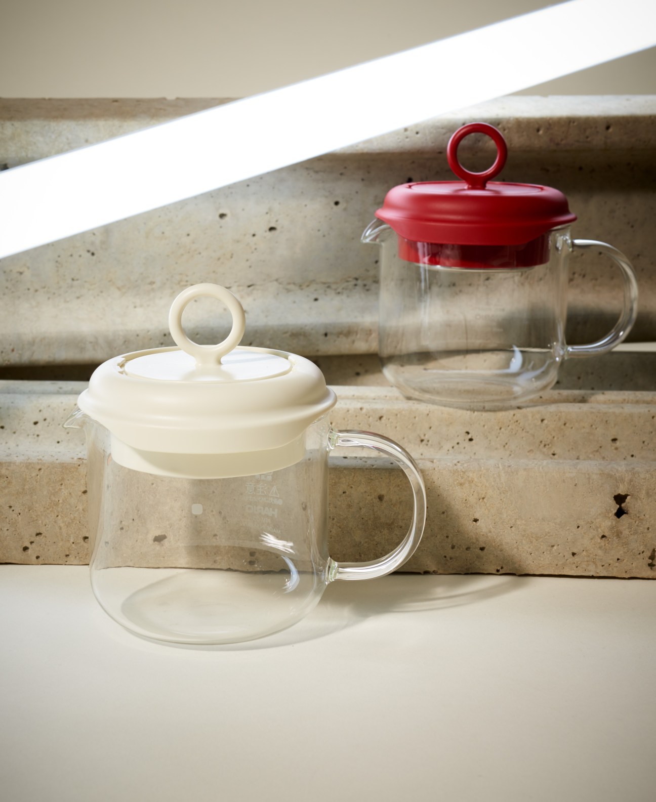 Two glass tea makers with built-in strainers by Hario, one with cream-colored lid and one with red lid.
