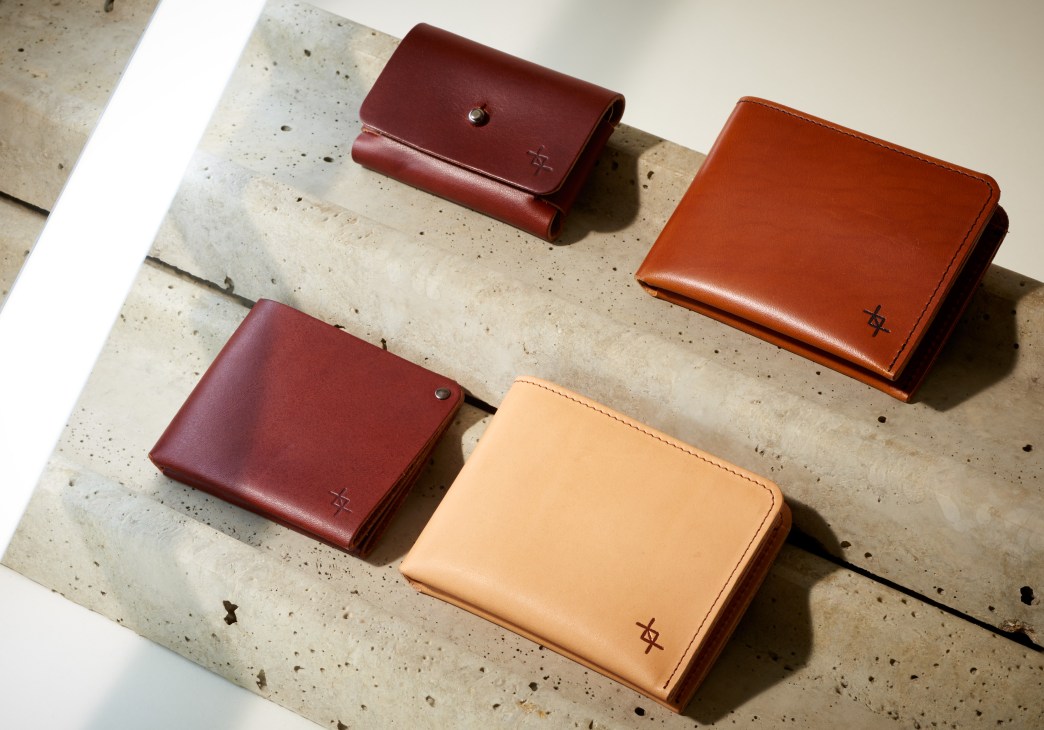 Four vegetable-tanned leather wallets and pouches by The Craftory in burgundy, rust, and tan colors arranged on a concrete surface.