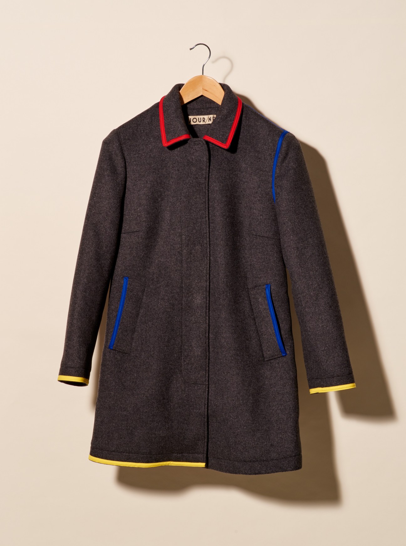 Dark grey wool coat by Jour/né with red collar and blue and yellow trim details on a wooden hanger.