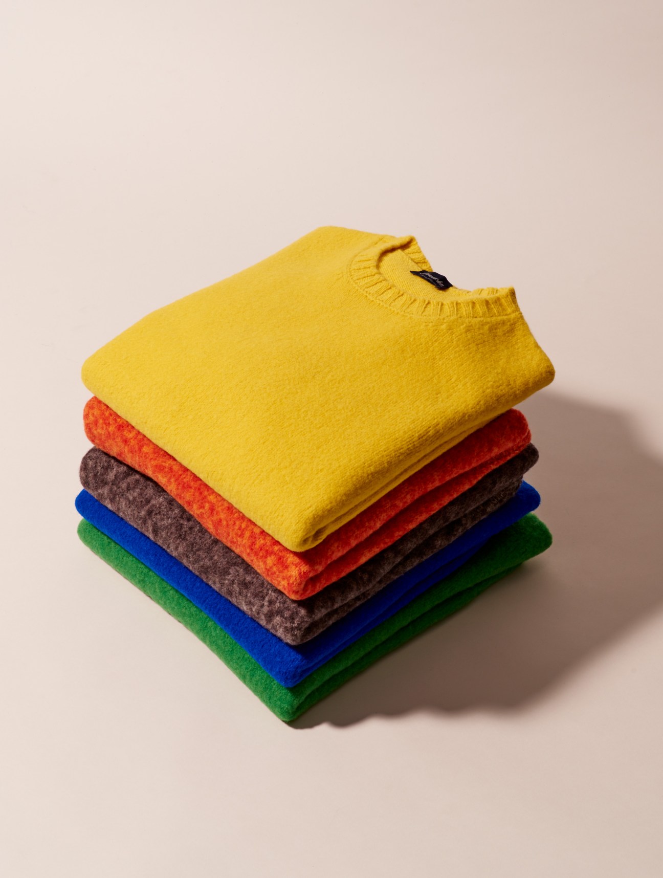 Stack of Drumohr knit jumpers in 11 different colors including yellow, red, orange, brown, blue and green.