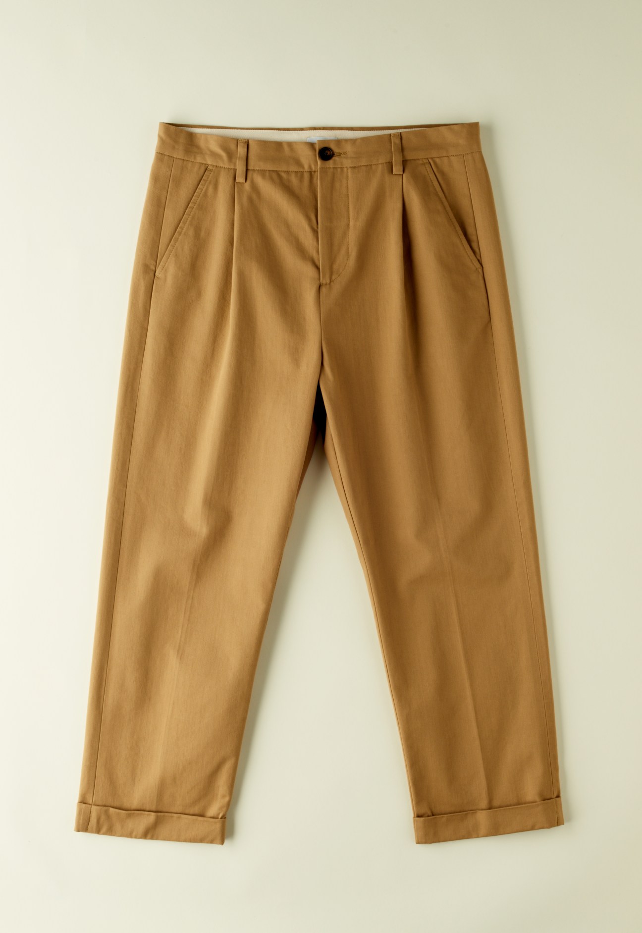 Tan pleated chinos with cuffed ankles displayed flat against a light beige background.