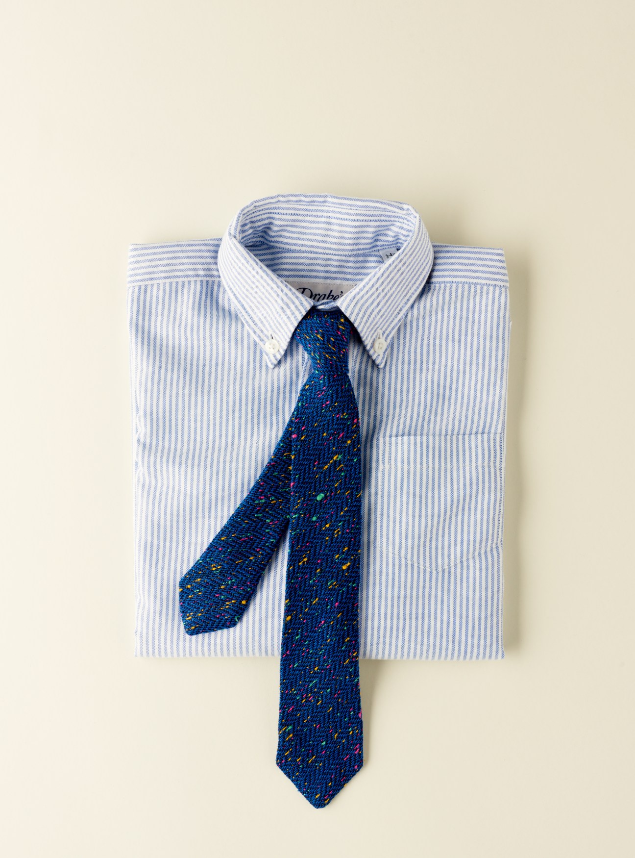 Drake's blue patterned tie displayed on a light blue striped dress shirt against a cream background.