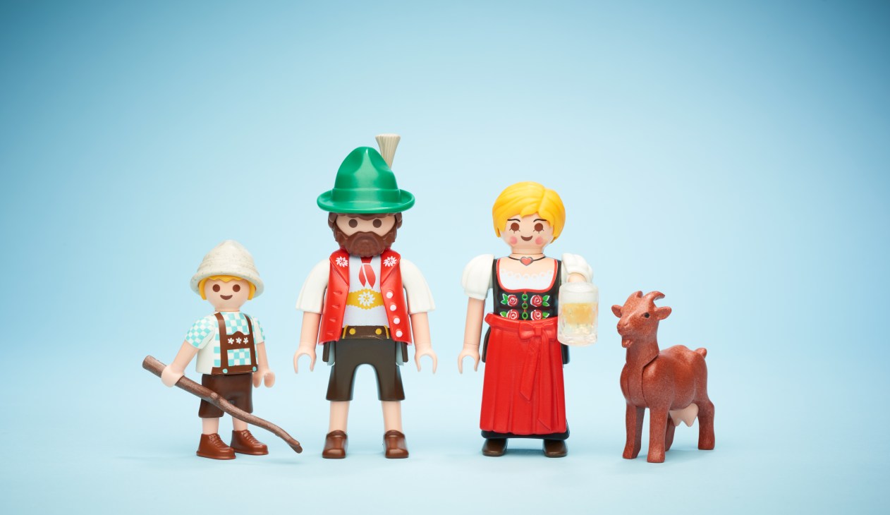Playmobil figures dressed in traditional Alpine tracht clothing including lederhosen, dirndl, and hat against a blue background.