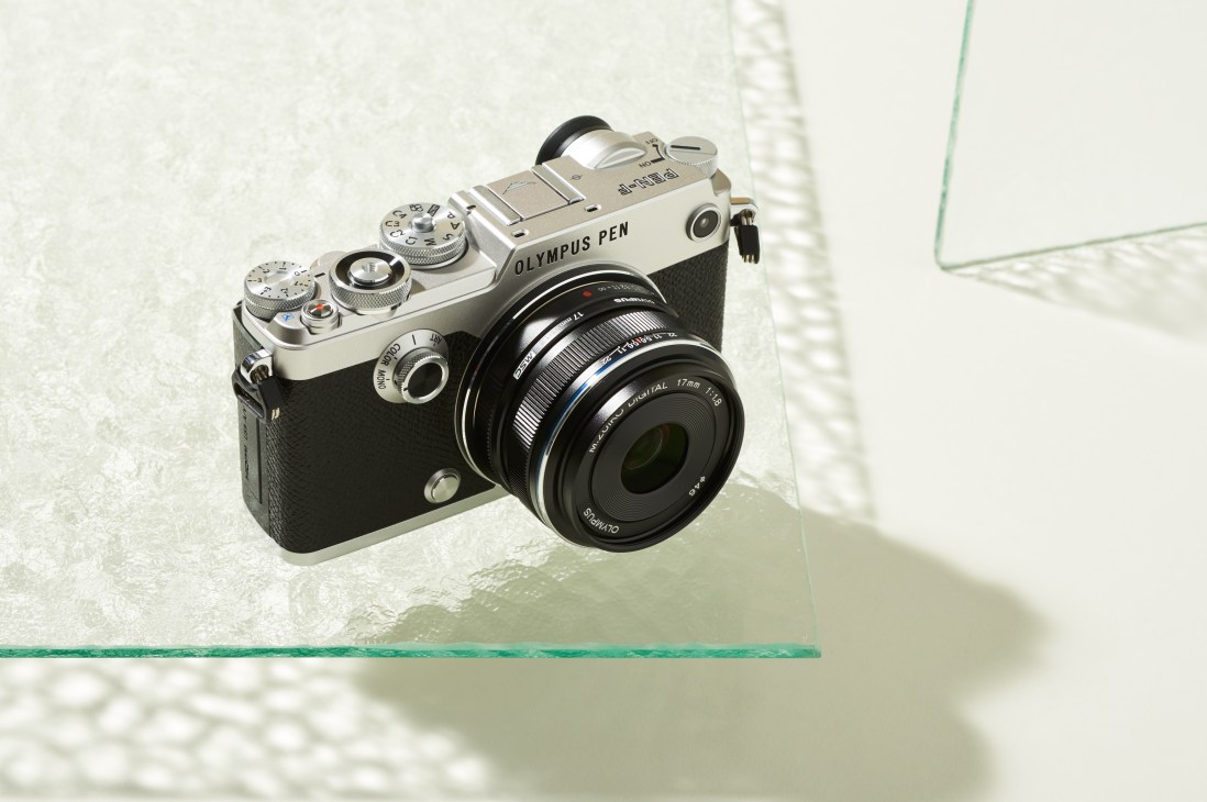 Olympus PEN-F camera with silver and black body displayed on a light surface with shadow.