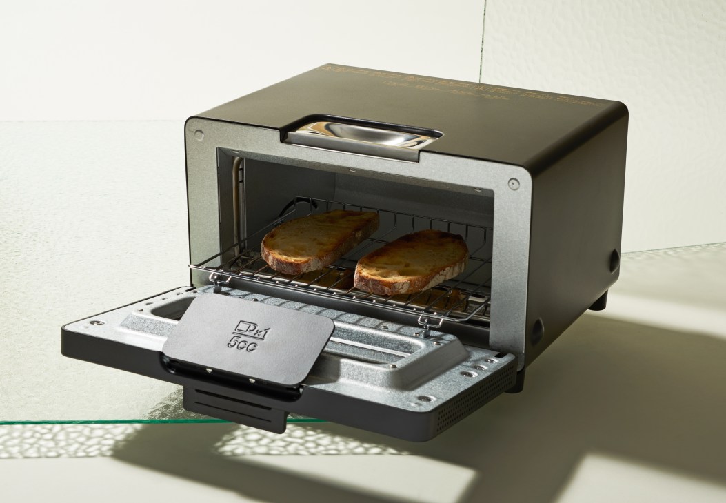 Balmuda toaster with two slices of golden-brown toast inside, featuring a steam misting function and pull-out crumb tray.