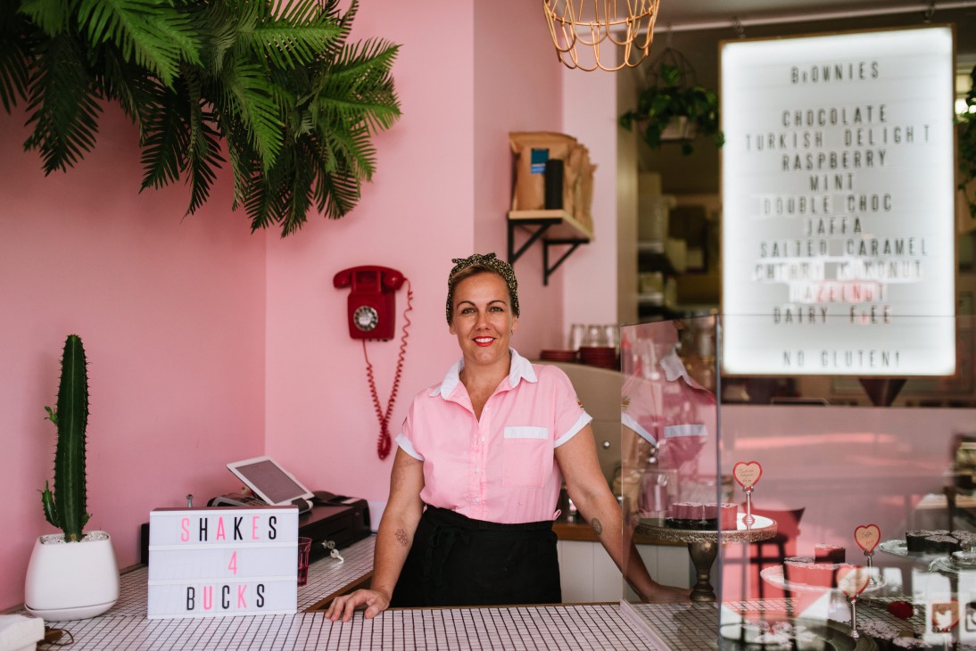 Lulu Purnell stands behind the pink counter of I Heart Brownies café, smiling at the camera in her pink shirt.