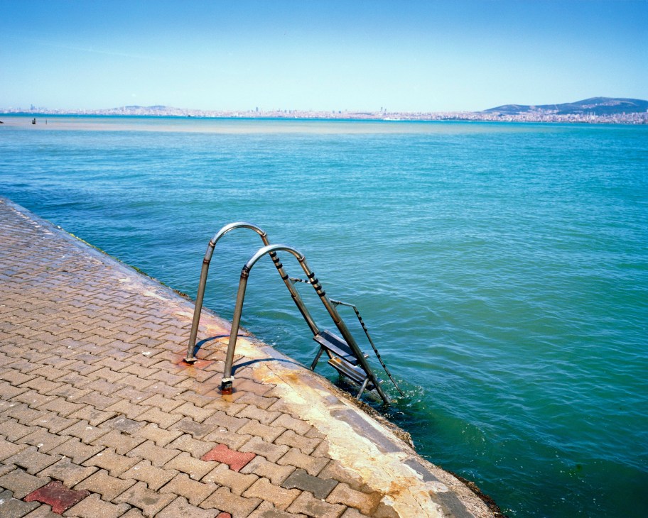 Metal ladder steps leading into turquoise water on a paved waterfront with Istanbul skyline visible across the Bosphorus.