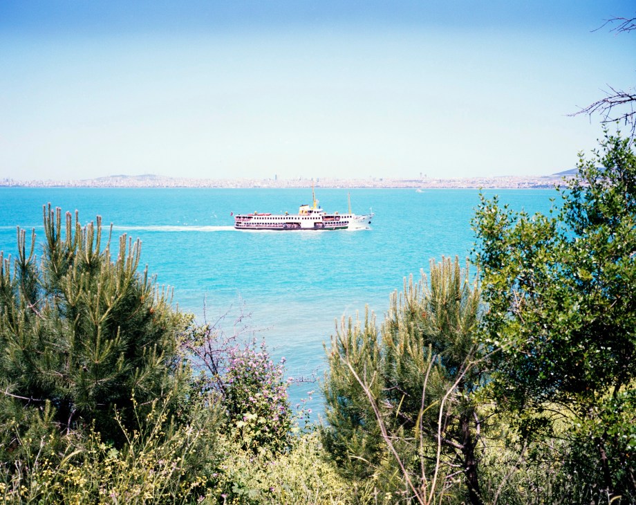 A passenger ferry travels across turquoise Bosphorus waters toward distant Istanbul skyline, framed by Mediterranean vegetation.