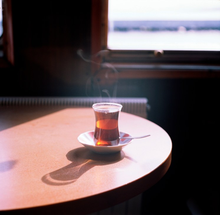 Turkish tea in a traditional glass on a white saucer with a spoon, placed on a round table with the Bosphorus visible in the background.