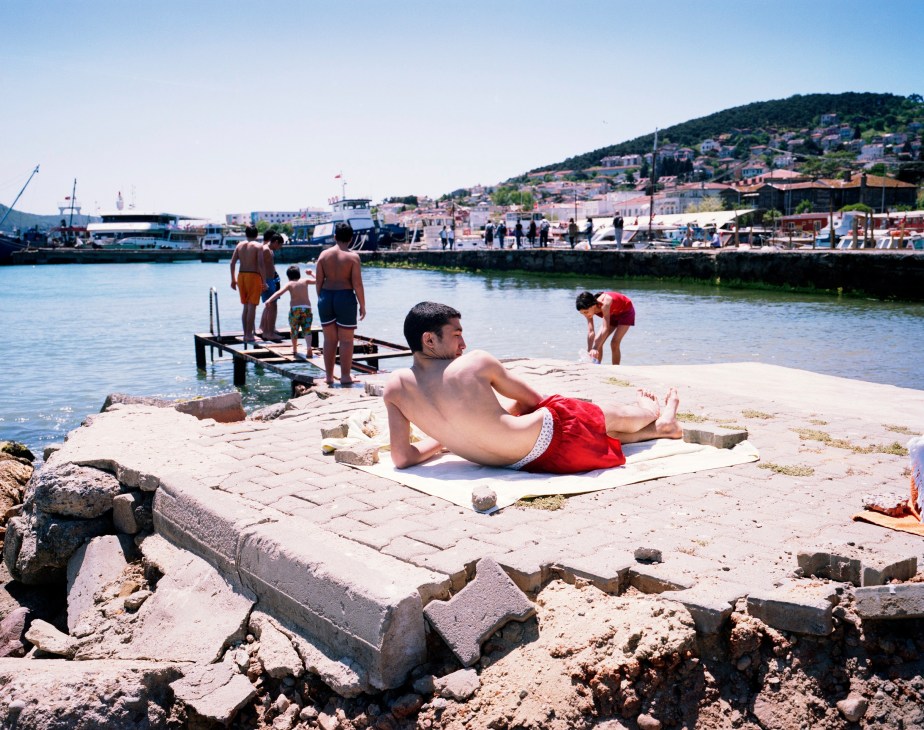 Bathers relax and swim at a rocky waterfront on Heybeliada in the Princes' Islands, with ferries and hillside buildings visible across the water.