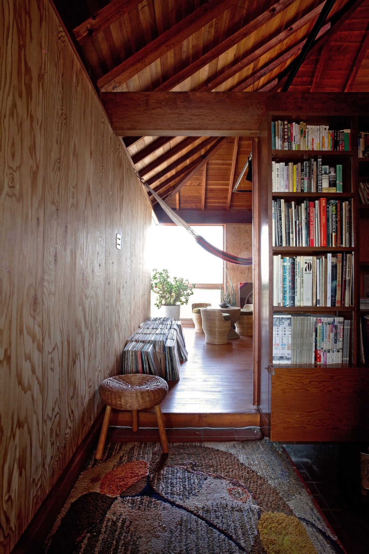 Wood-panelled hallway with exposed beam ceiling, vinyl records stacked on floor, woven rattan stool, and built-in bookshelf with multicolored books.