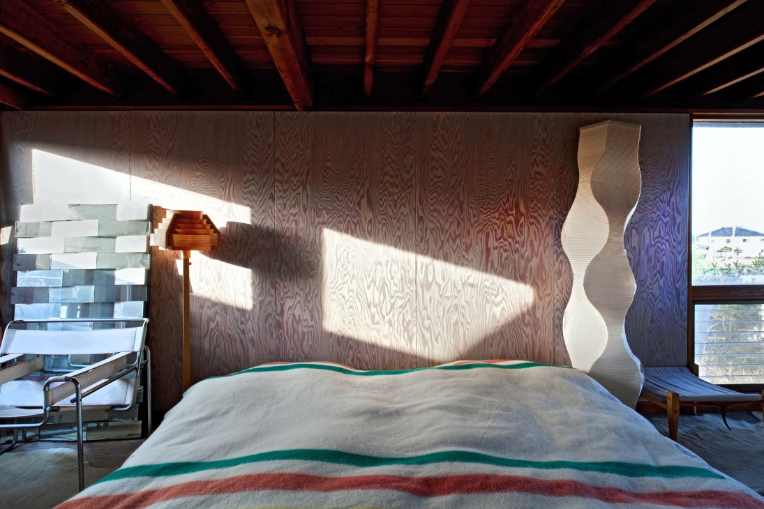 Sunlit bedroom with wooden walls, exposed beam ceiling, handmade bed with striped blanket, and modern sculptural wall art.