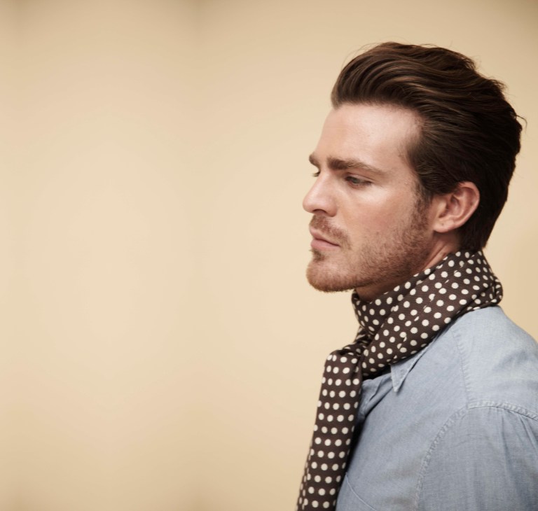 Man wearing Jane Carr polka dot scarf and light blue shirt against cream background.