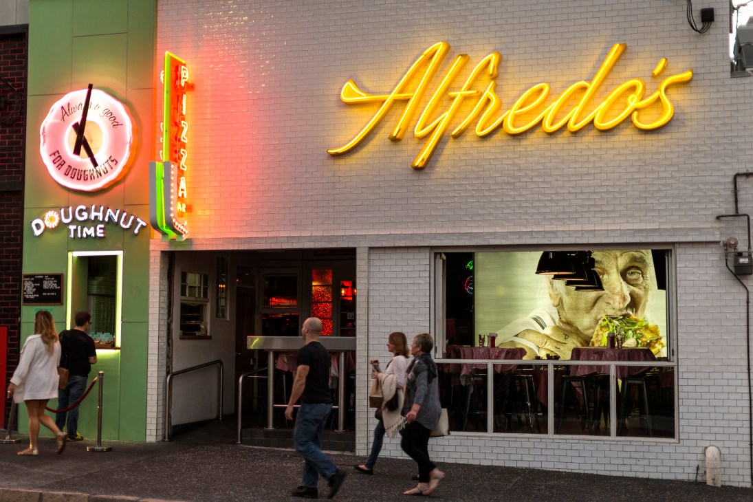 Alfredo's Pizzeria storefront in Brisbane with yellow neon sign and people outside.
