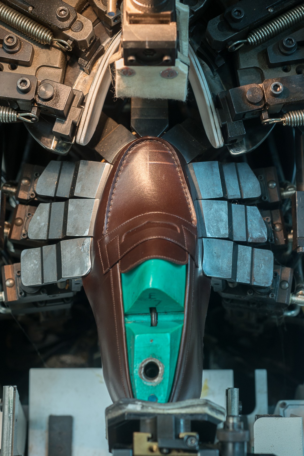 A brown leather shoe being assembled in JM Weston's machine with mechanical clamps and a turquoise base.