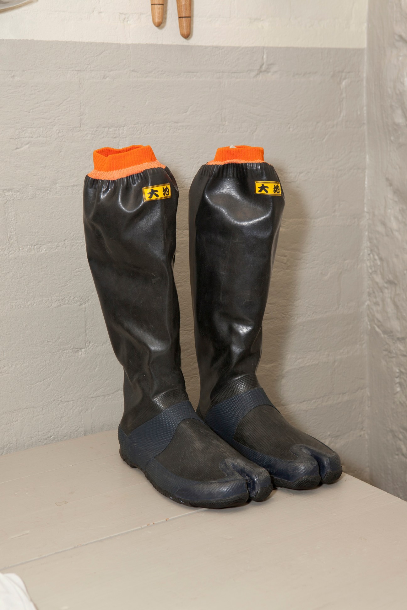 Pair of black rubber boots with orange ribbed cuffs and yellow labels displayed on white surface.