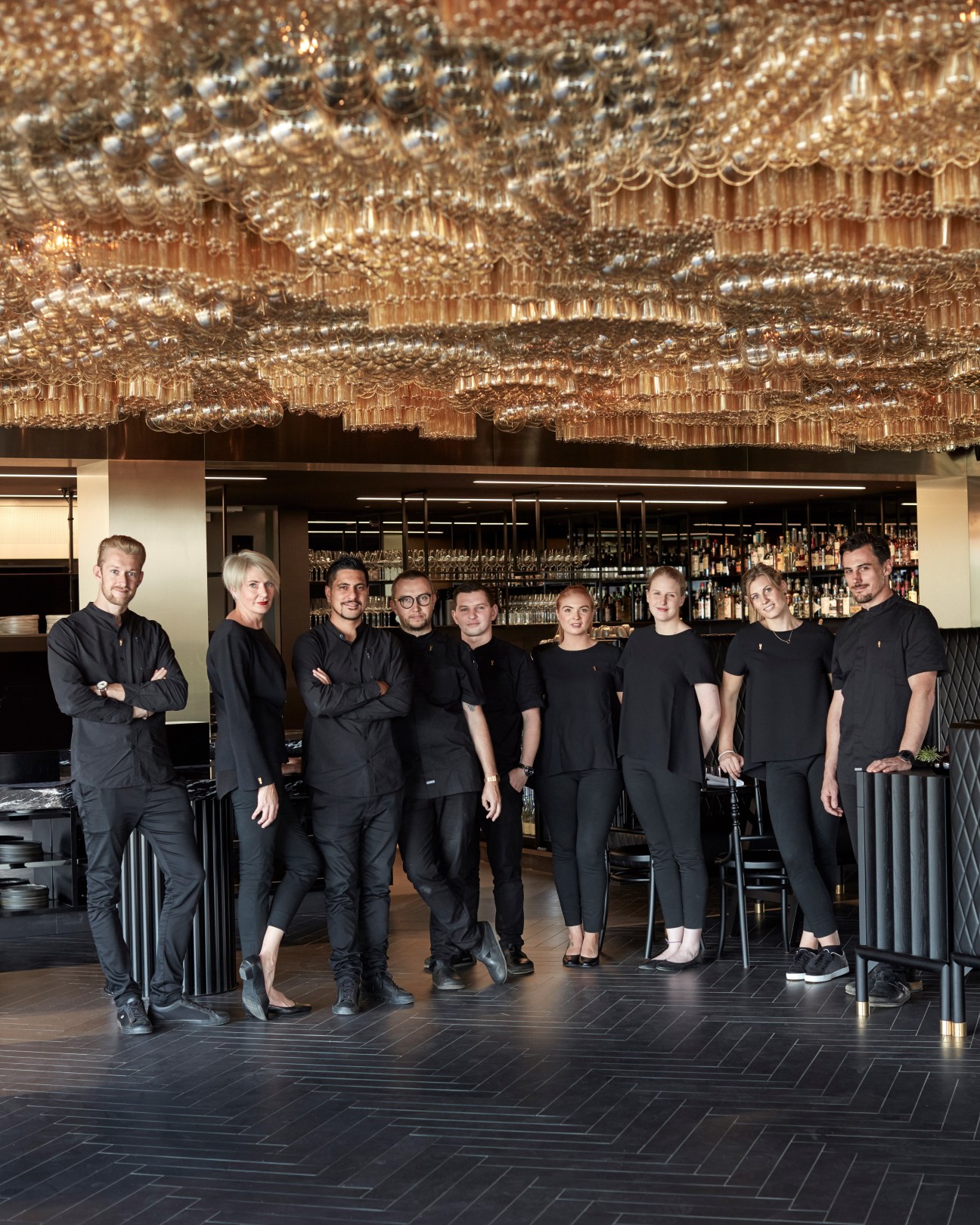 Ten hotel staff members in black uniforms stand together in the Jackalope Hotel bar under an ornate gold sculptural ceiling.