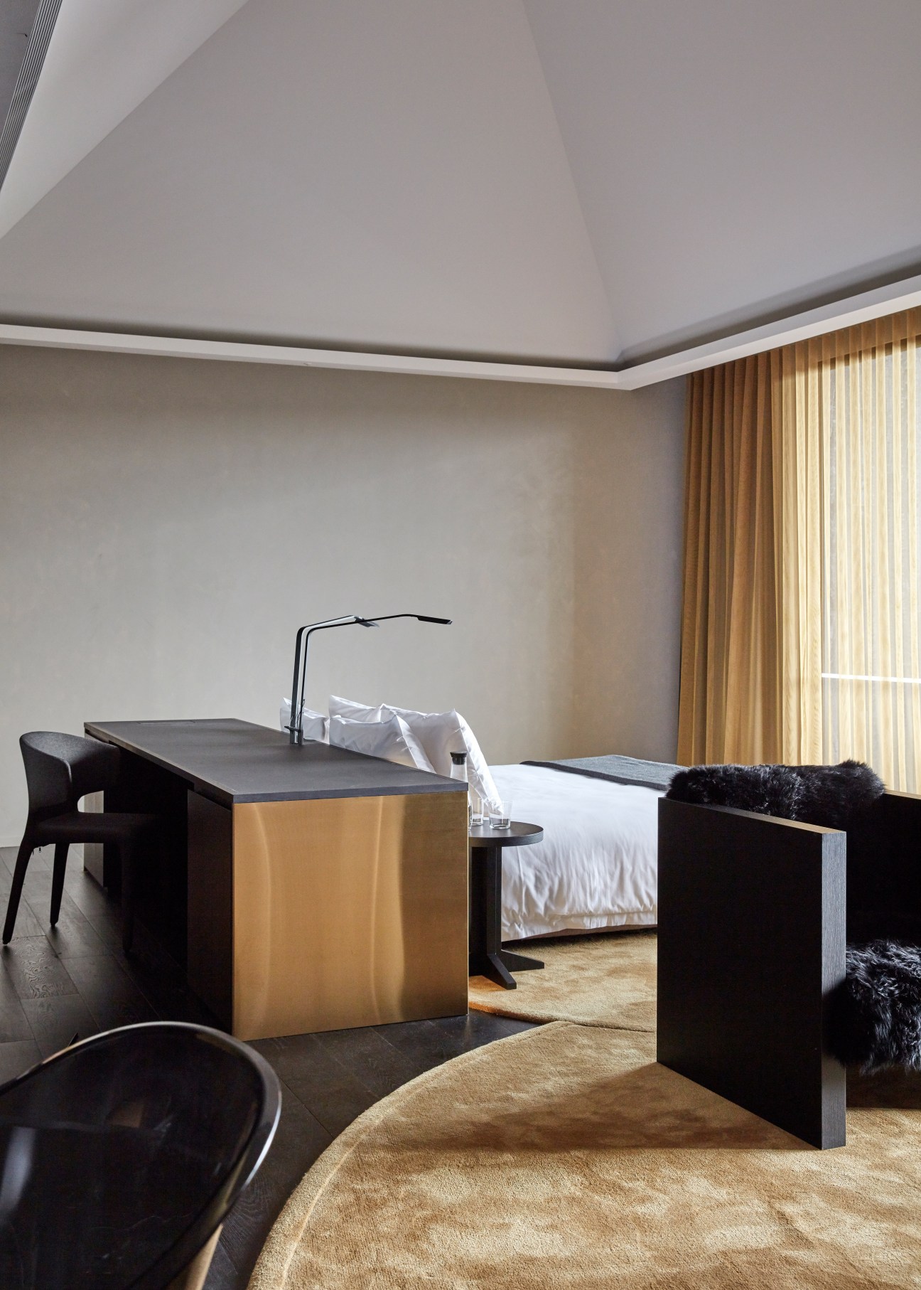 Minimalist hotel suite with gold desk, black chair, bed with white linens, and gold curtains overlooking vineyards.