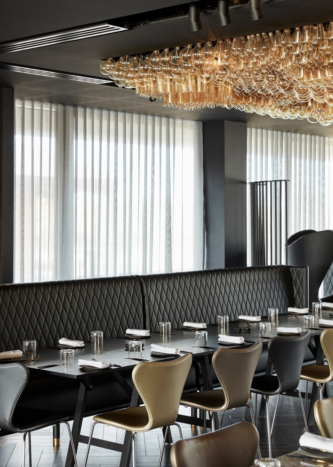Modern dining room with quilted black banquette seating, gold chairs, and ornate glass chandelier at Doot Doot Doot restaurant.