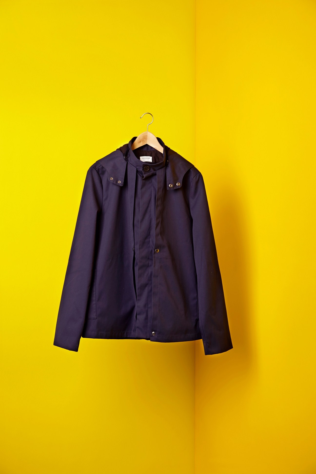 Navy waxed twill parka by 13 Bonaparte on wooden hanger against yellow background.