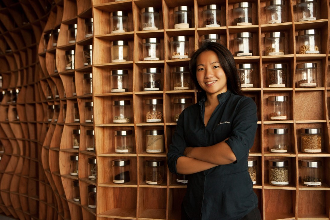 Janice Wong, pastry chef and founder of 2am:lab, poses in front of a wooden wall of shelves filled with glass jars of ingredients.