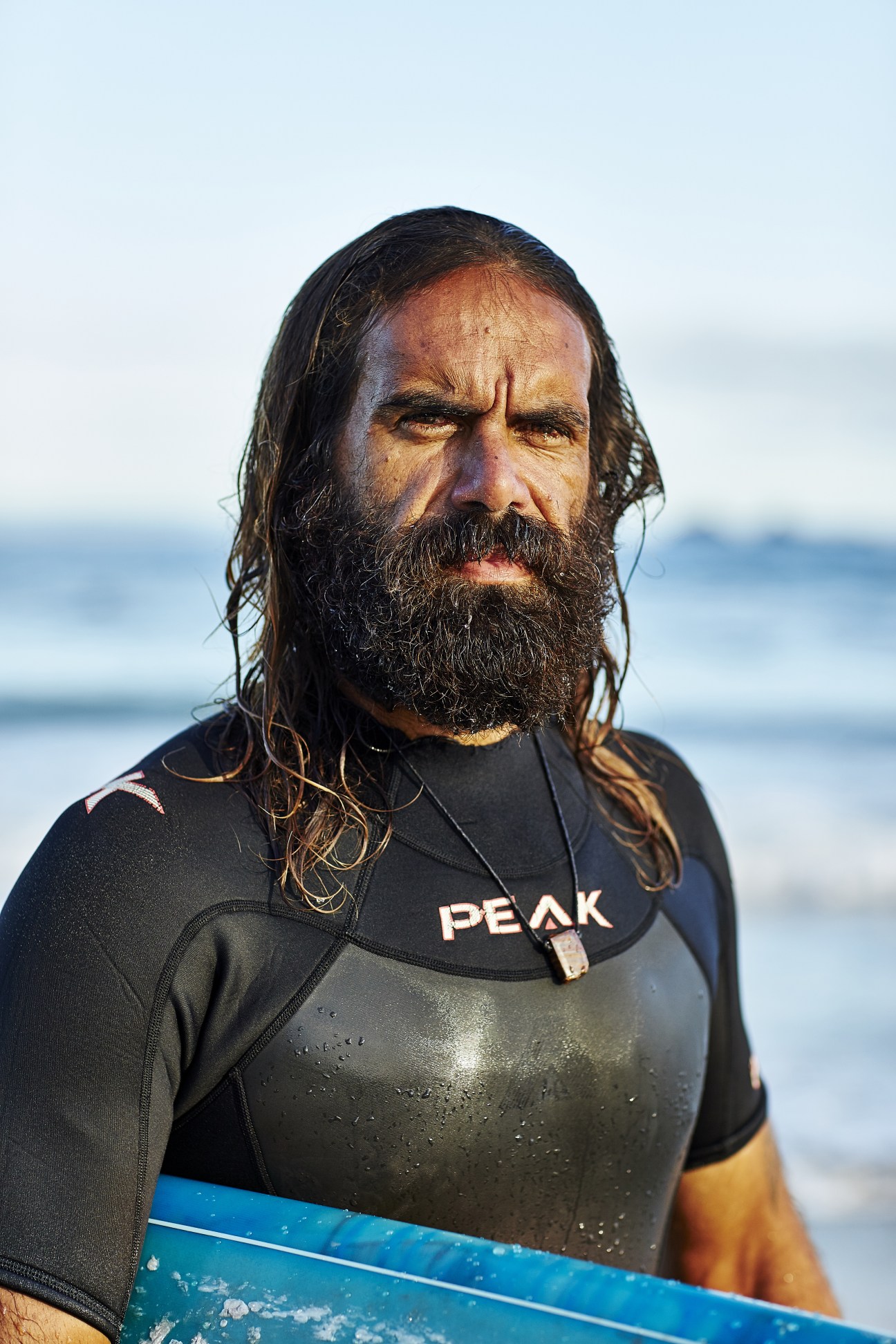 Bearded man in black wetsuit holding surfboard with ocean waves behind him