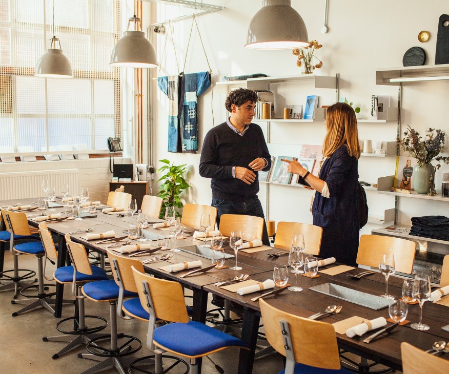 Two people in a bright factory space with wooden chairs and dining tables set for a meal, hanging denim samples visible above.