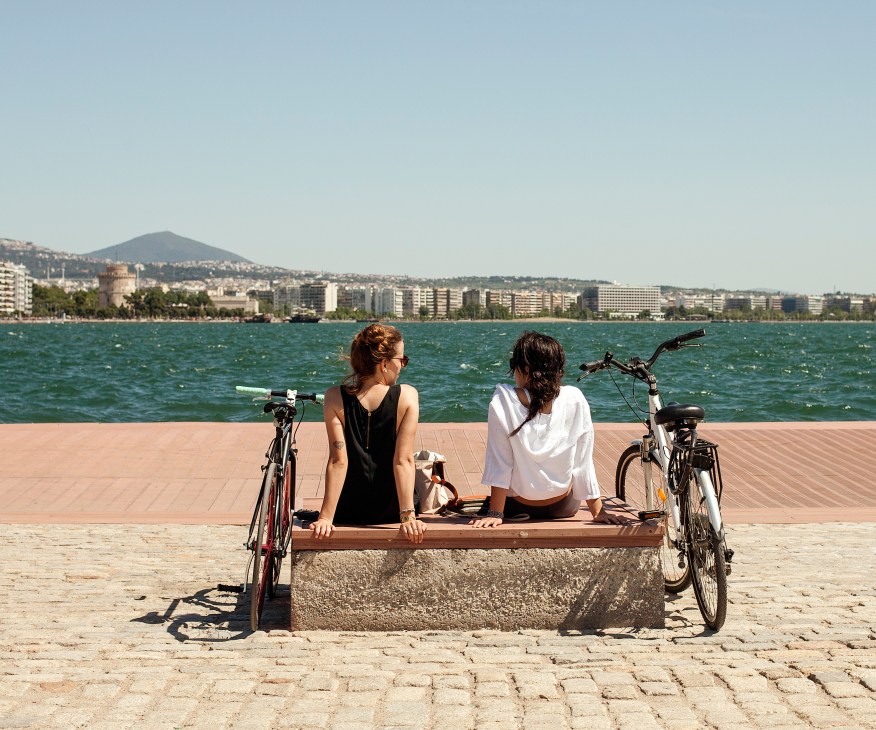 Two women sit on a wooden bench between bicycles on a sandy beach facing the sea and Thessaloniki's waterfront.