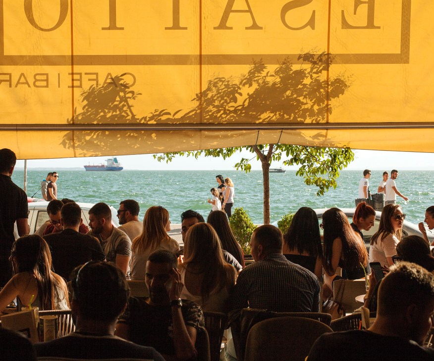 Crowded seaside café in Thessaloniki with yellow awning overlooking the Thermaic Gulf and moored ships.