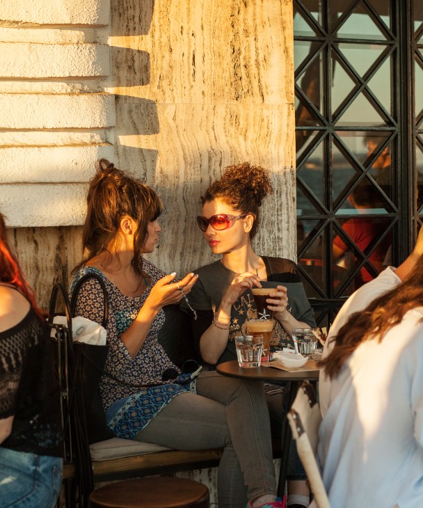 Two women sitting at an outdoor café in golden evening light, one holding a drink.