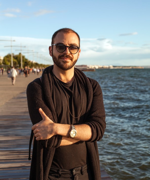 Man wearing sunglasses and dark cardigan standing on Thessaloniki seafront with water and pedestrians behind