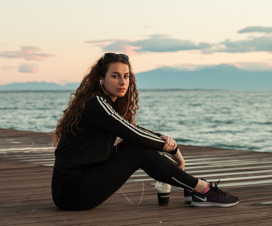 Woman in black athletic wear sitting on wooden deck by sea at sunset with coffee drink and earbuds.