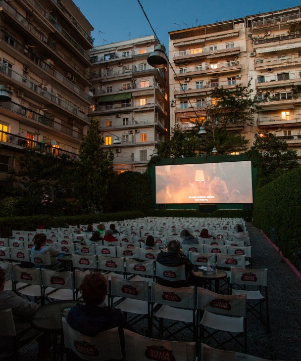 Audience sitting in white chairs at Natali open-air cinema in Thessaloniki with a film projected on screen, surrounded by apartment buildings at dusk.