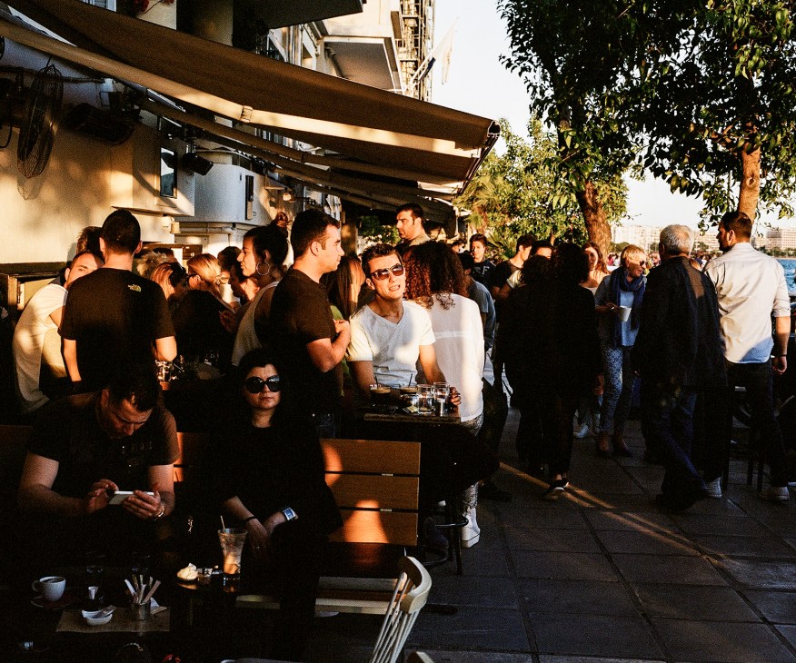 Crowd of people gathered at an outdoor café with a retractable awning on a sunny day in Thessaloniki.