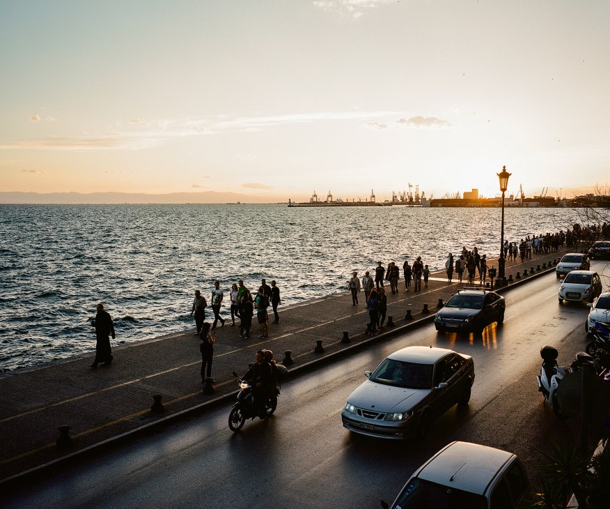 People gather on Thessaloniki's waterfront esplanade at dusk with cars parked along the road and boats visible in the harbor.