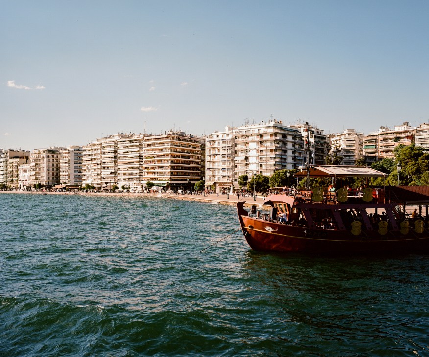 A wooden floating bar anchored in the sea with Thessaloniki's seafront buildings in the background.