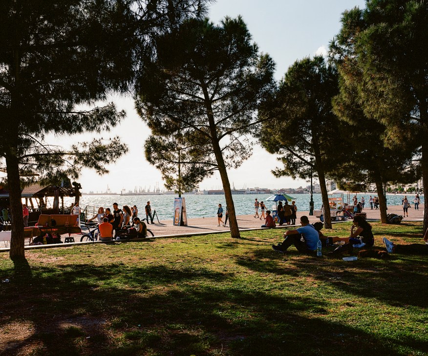 People relaxing under pine trees in a grassy park overlooking the sea and city skyline in Thessaloniki.