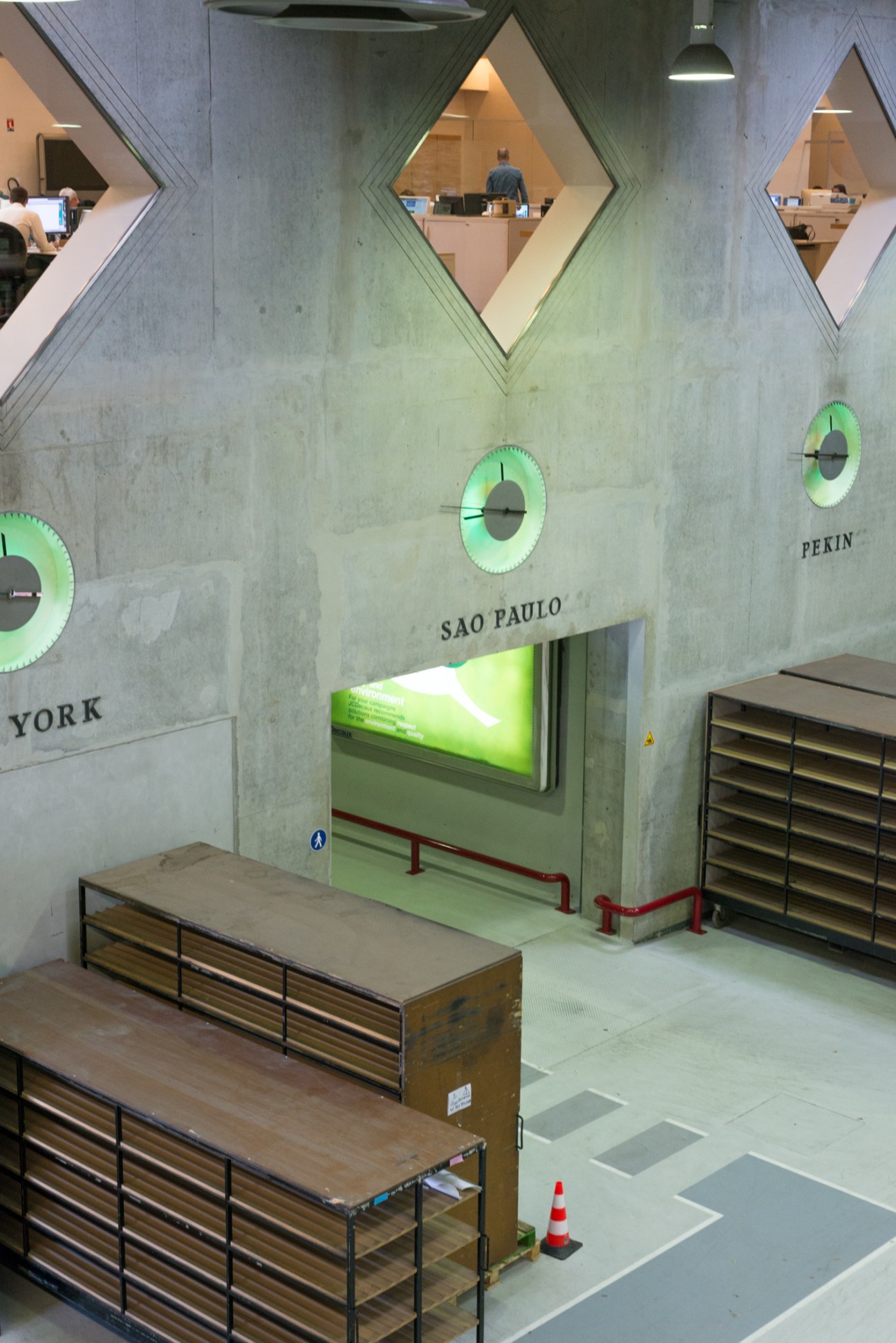 Interior of JCDecaux printing facility with concrete walls, illuminated clocks labeled with city names, and wooden printing equipment.