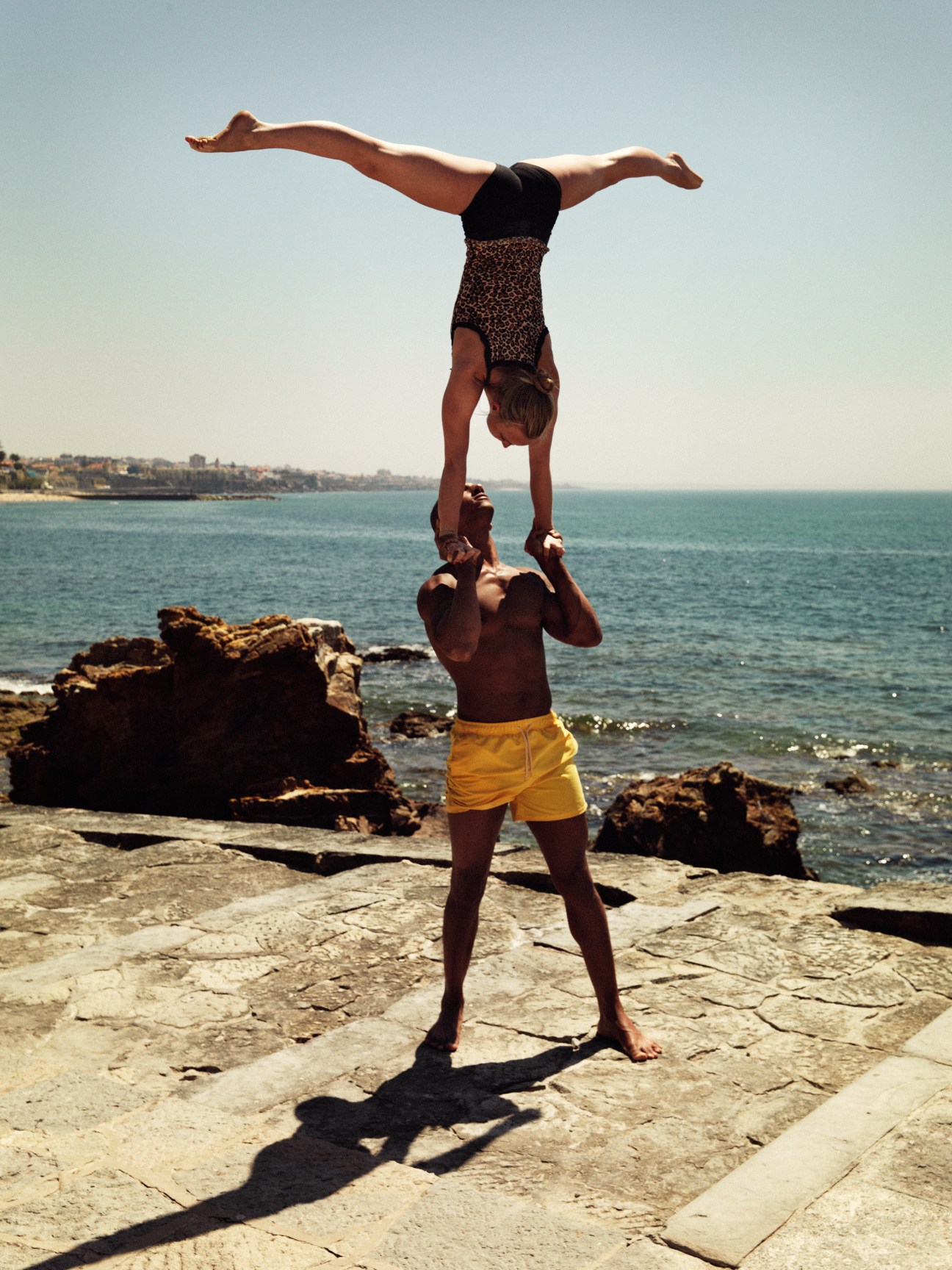 Couple performing an acro yoga handstand on rocky coastal promenade with sea and beach in background.
