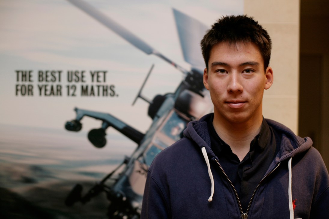 Young man in dark hoodie standing in front of military recruitment poster with helicopter imagery.