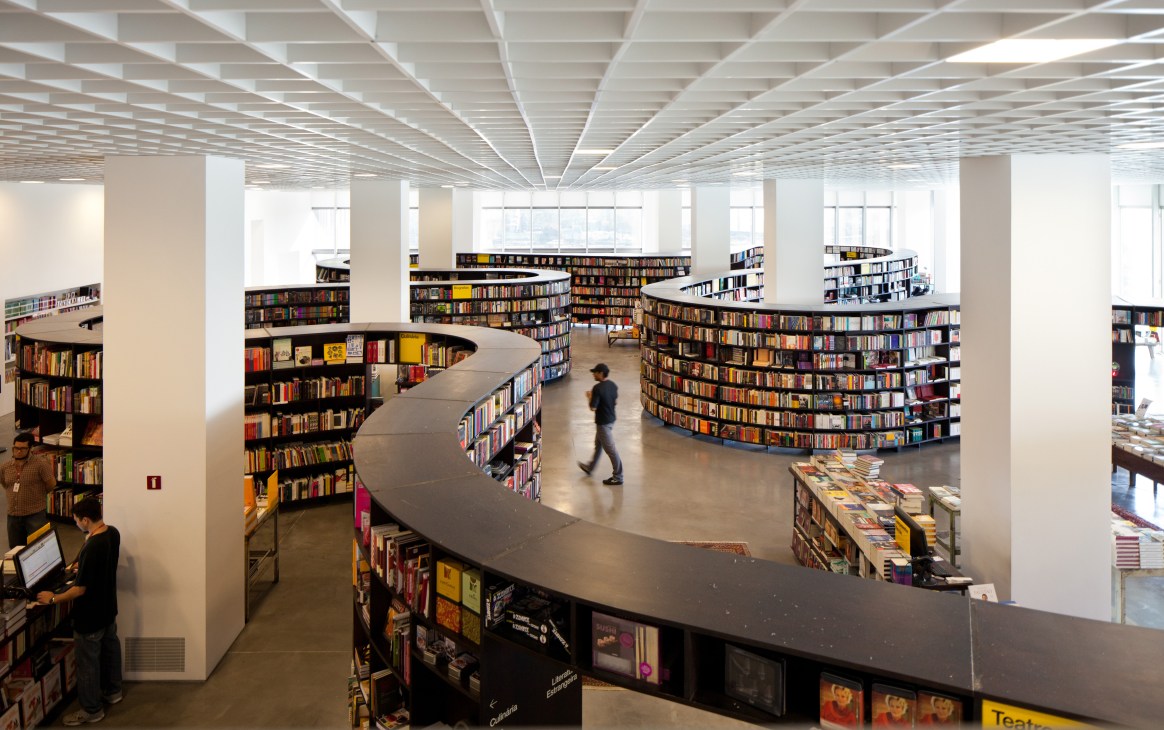 Interior of JK Iguatemi mall with curved bookshelf displays, white ceiling, and person walking through contemporary retail space.
