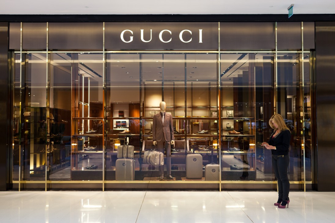 Woman browsing outside a Gucci storefront displaying menswear and accessories in a modern mall setting.