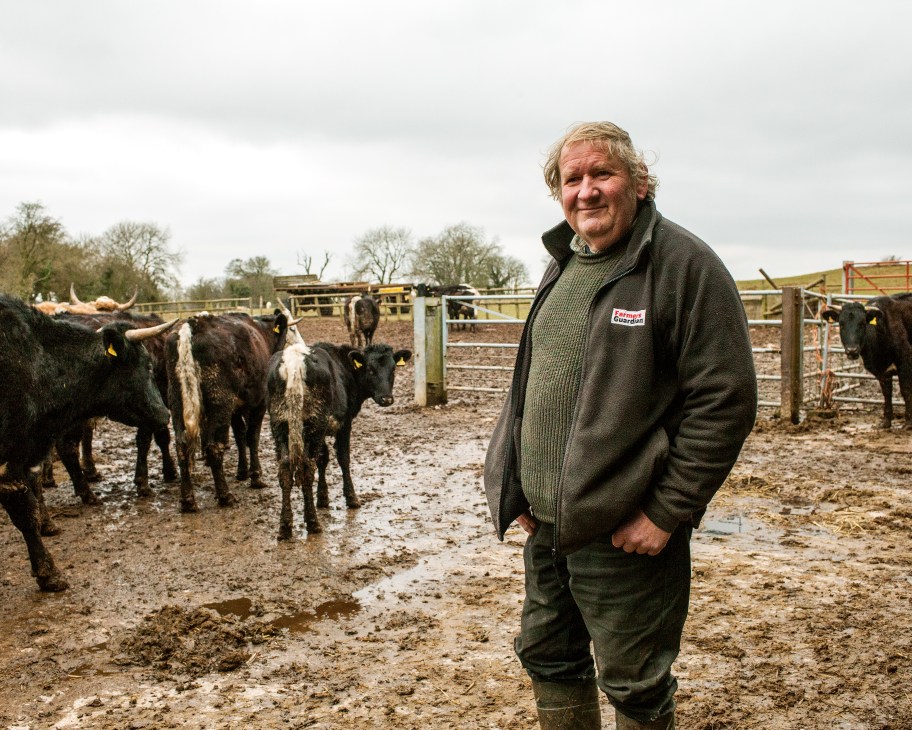 Farmer John Childs standing in a muddy farmyard with rare-breed cattle behind him.