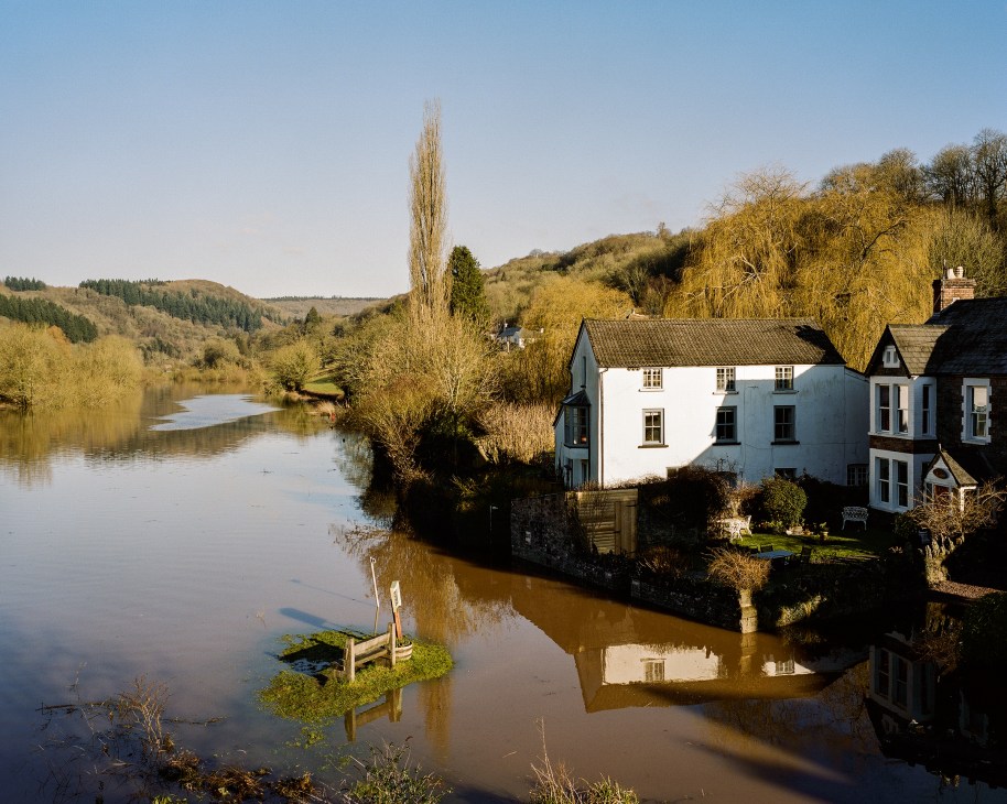 White cottage beside the Wye River with wooded hills reflected in calm water.