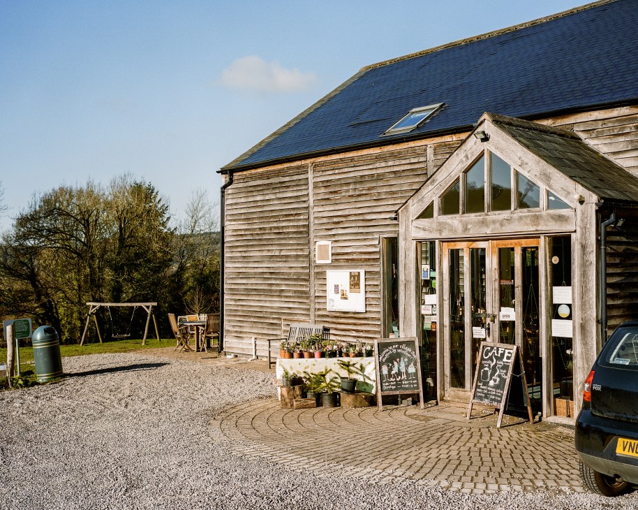 Exterior of Brockweir and Hewelsfield Village Shop, a contemporary wooden building with green oak construction, featuring café signage and potted plants at the entrance.