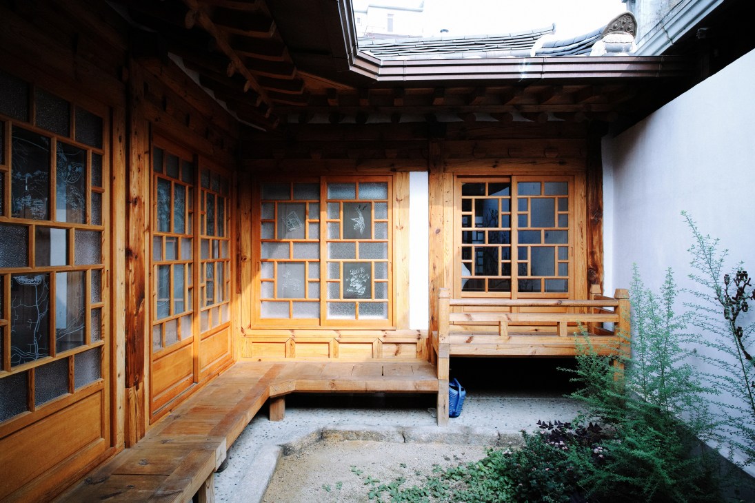 Traditional Korean hanok courtyard with wooden walkway, gridded windows, and white walls under a skylight.