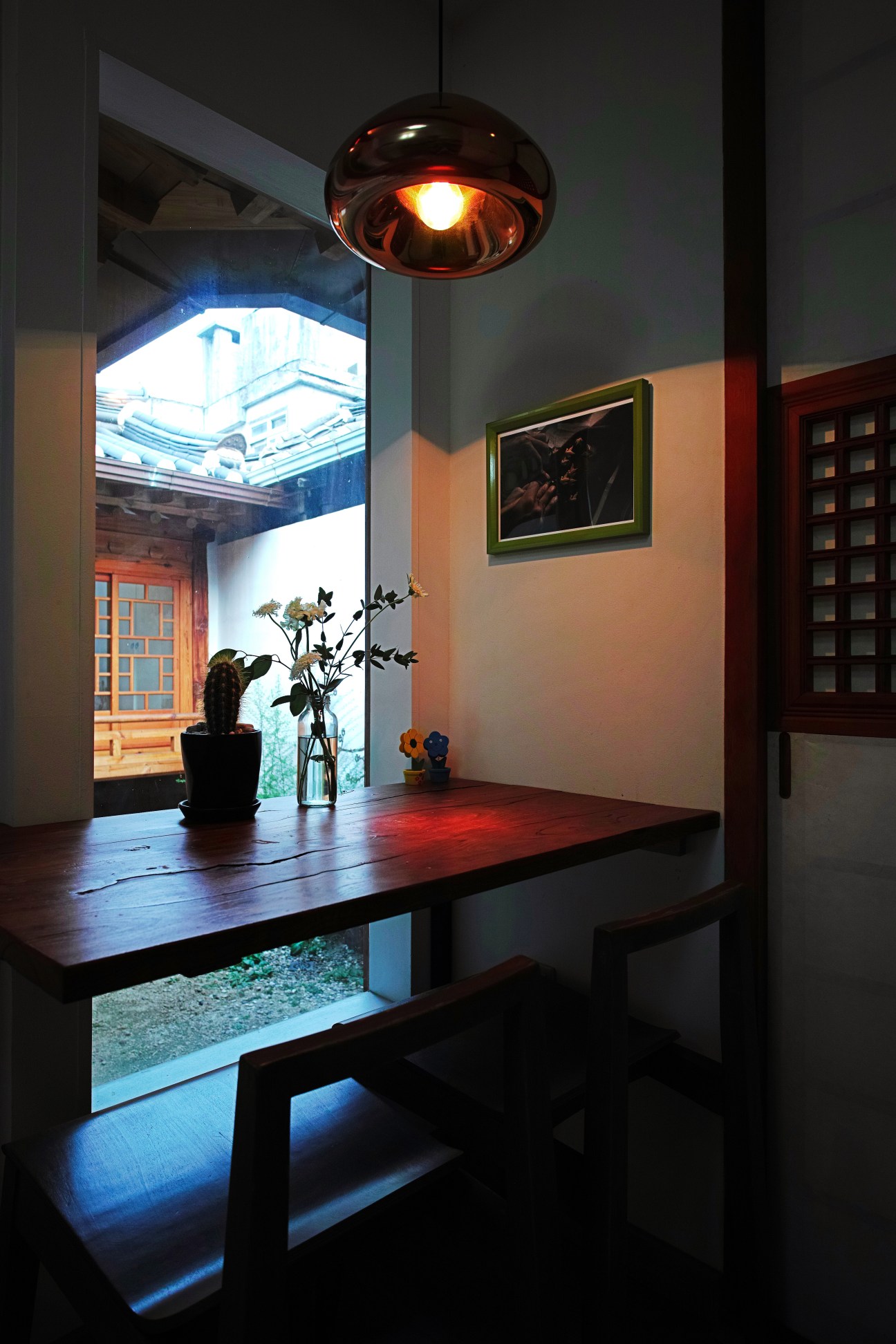 Modern dining area in restored hanok with wooden table, pendant lamp, potted plant and view to courtyard.